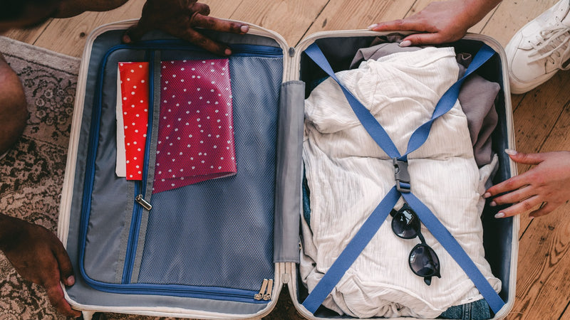 Friends packing an open suitcase on the floor with a light summer top, denim and sunglasses, white trainers beside it